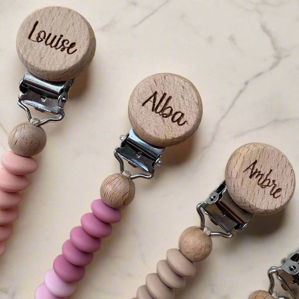 Three wooden pacifier clips with pink and beige beads on a light background
